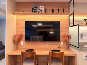 A modern kitchen with a large flat-screen TV, wooden cabinetry, and a dining area with two chairs. - Condomínio Helbor Edition  (São Paulo)