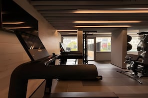 Ginásio com esteira, aparelhos de musculação e uma grande janela com vista para edifícios.