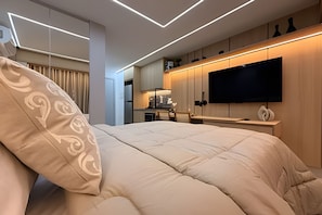 Quarto de hotel moderno com uma cama grande, televisão de tela plana na parede e uma área de kitchenette.