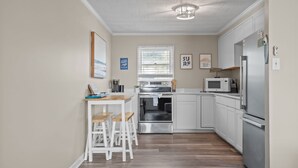 Una cocina moderna con muebles blancos, un refrigerador de acero inoxidable, un microondas y un horno doble. Hay dos taburetes de madera junto a una pequeña mesa, y una ventana con persianas.