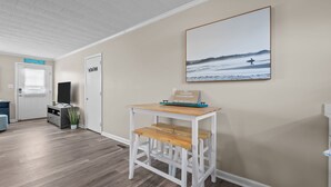Una habitación con una mesa de madera, un banco blanco, una puerta, un cuadro colgado en la pared y un letrero en la pared.