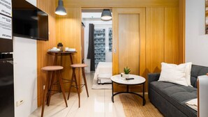 A compact living space with a sofa, a small round table, a TV, and a kitchenette area. - Brand New Luxe Japandi w Pool View MOA 15 min NAIA (Pasay City)