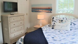 Une chambre à coucher avec une télévision posée sur une commode, une lampe de chevet, un lit recouvert d’une couvre-lit à motifs floraux et orné d’un coussin avec un texte, et un tableau représentant une jetée accroché au mur.