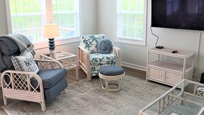 Une pièce comprenant un fauteuil en osier, un pouf, une petite table avec une lampe, un téléviseur fixé au mur et un meuble.