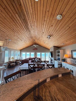 Un vaste espace de vie doté d’un haut plafond en bois, d’une table à manger et d’un coin cuisine.