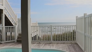 Una terraza de madera con vista al océano, una piscina y una escalera que lleva a una zona cubierta.