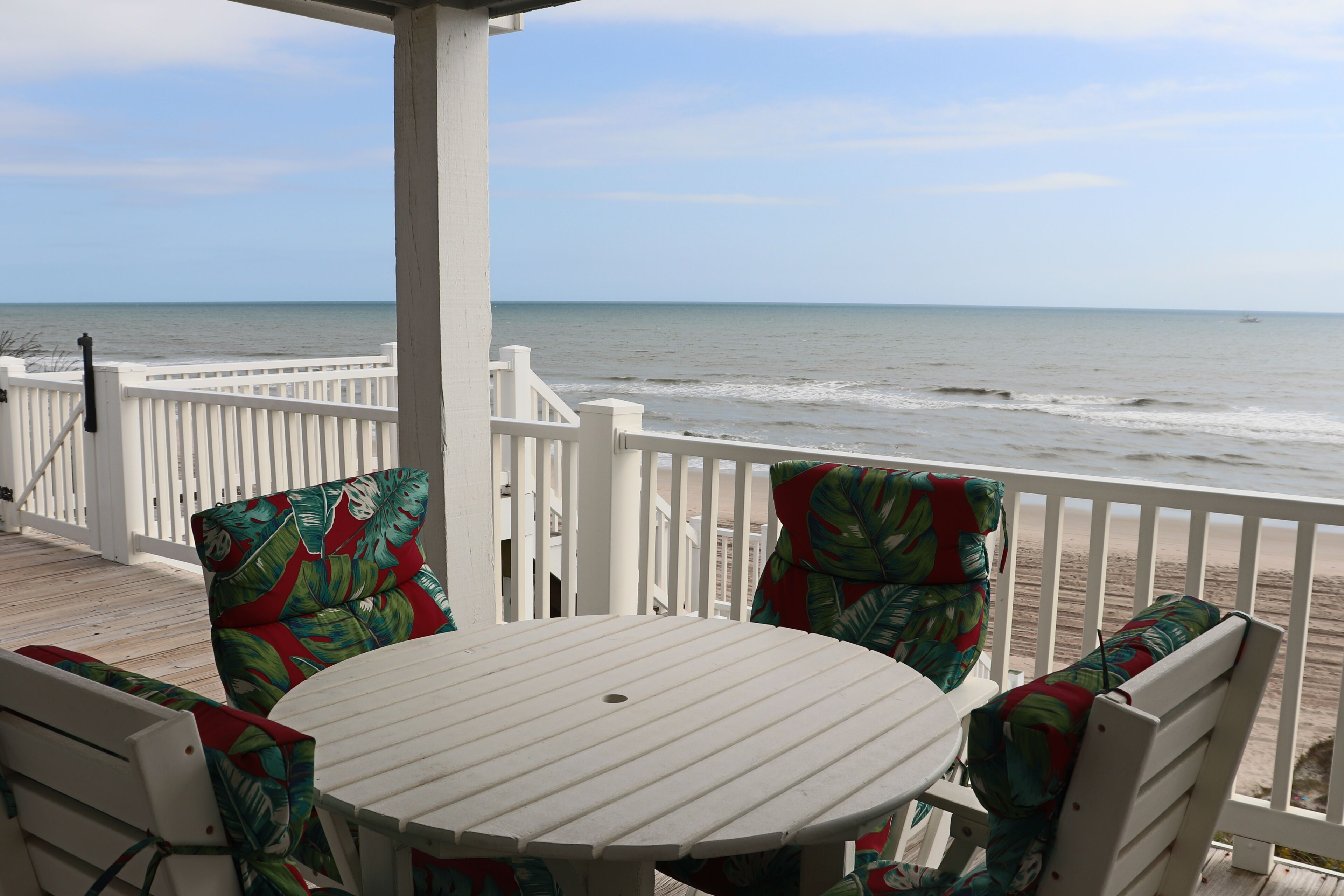 A white wooden deck with a round table and chairs, overlooking a beach.