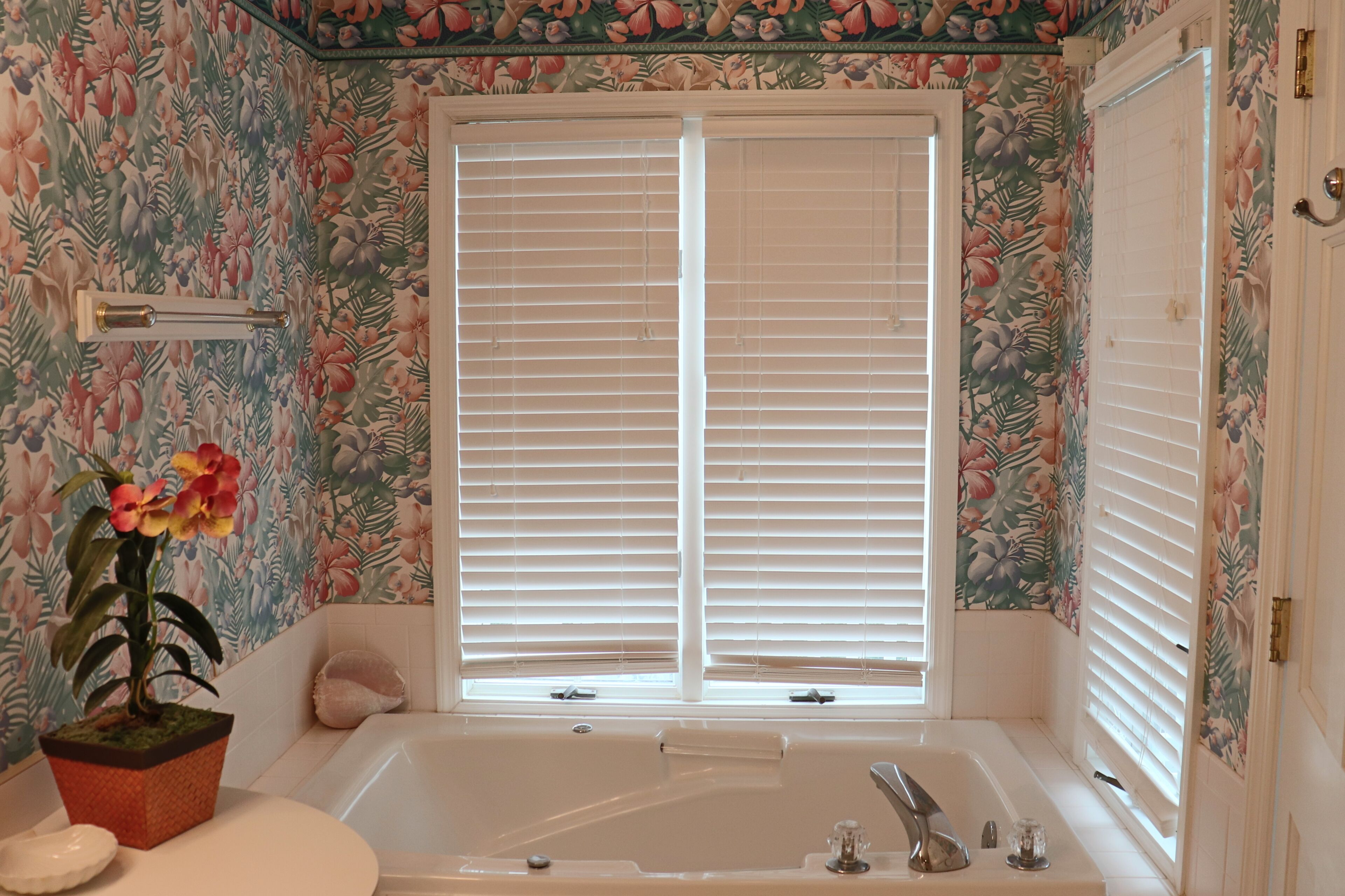 A bathroom with a bathtub, a window with blinds, a potted plant, and floral wallpaper.