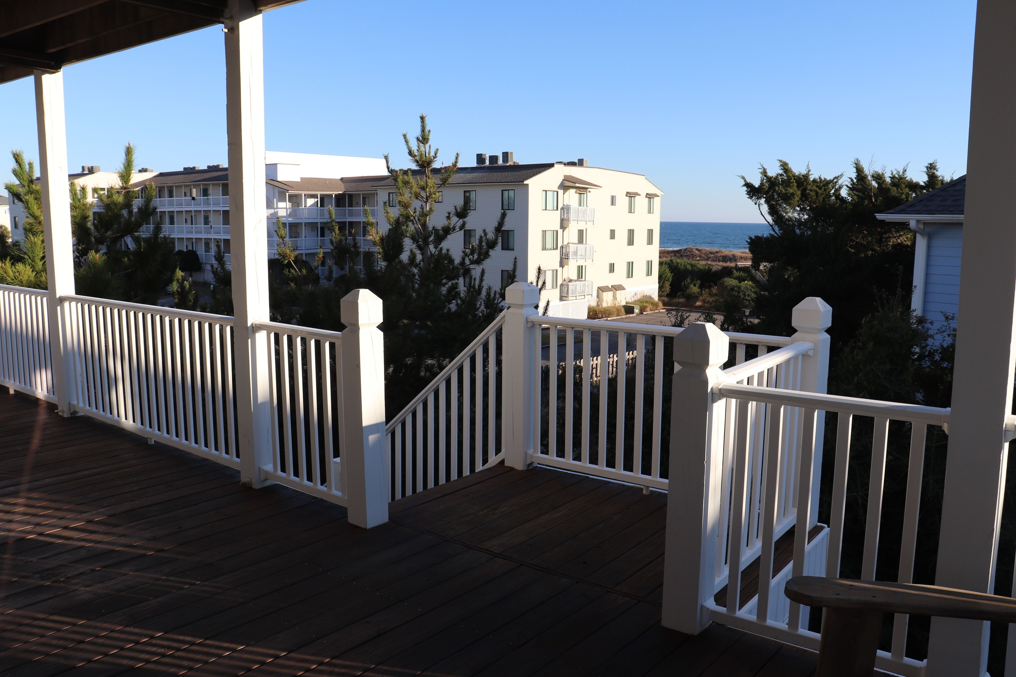 A wooden deck with white railings, overlooking a beachfront with buildings and trees.