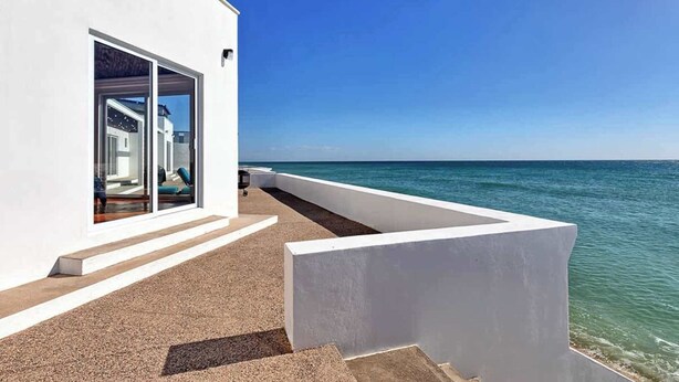 Um edifício branco moderno com porta de vidro que dá para um quarto, vista clara do oceano e uma estrutura de concreto em primeiro plano.