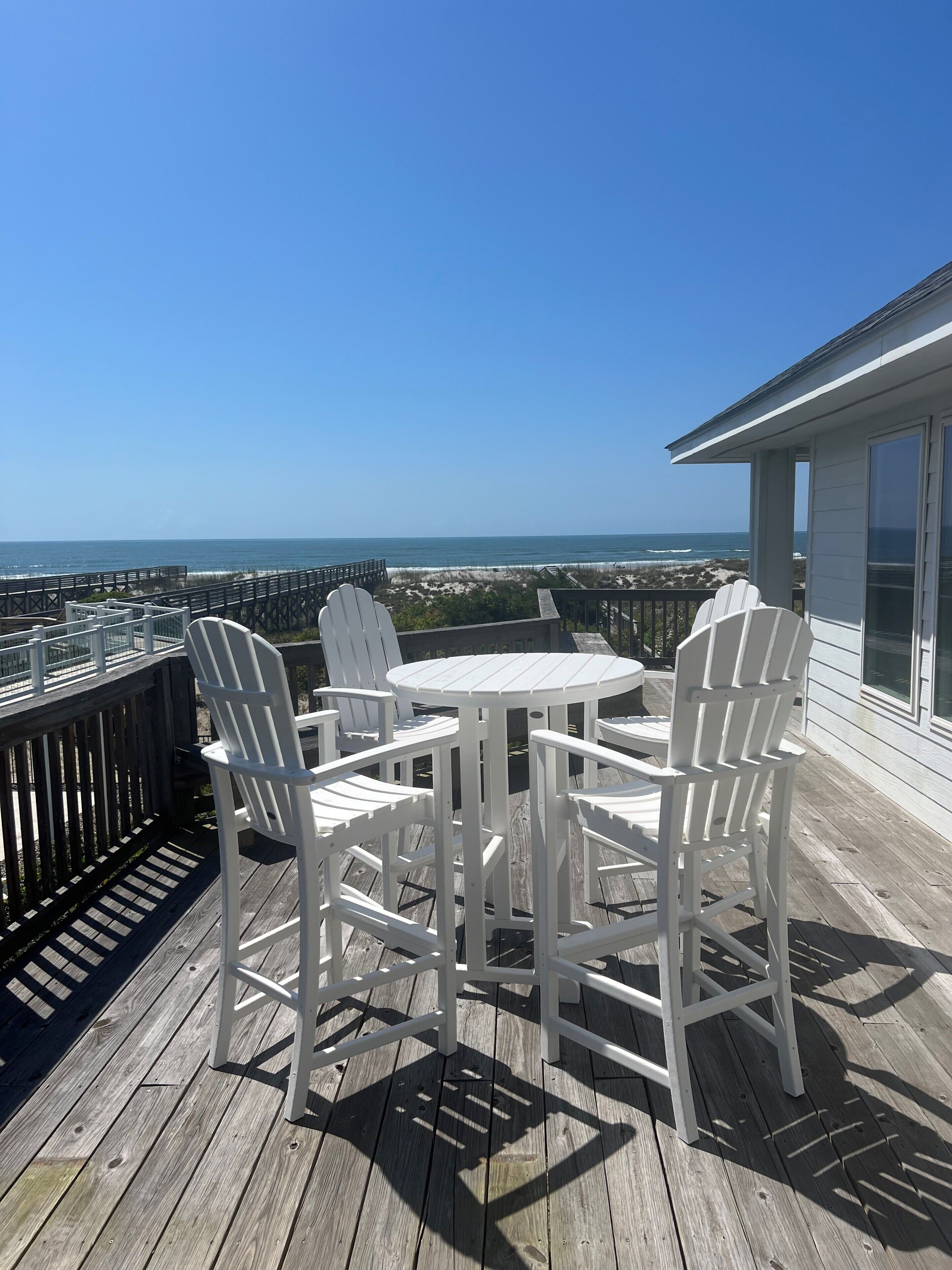 Eine Holzterrasse mit weißen Stühlen und einem Tisch, mit Blick auf Strand und Meer.