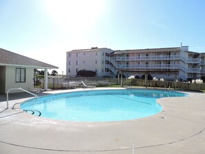 A swimming pool with a concrete deck, a wooden fence, and a multi-story building in the background. - Sunset Condo w/ Water Views & Pool Access (Emerald Isle)