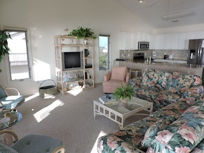 A living room with a sofa, a television on a stand, a coffee table, and a kitchen area with cabinets and appliances. - Oceanfront 4BR Home with Decks & Beach Access (Indian Beach)