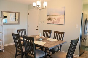 A dining area with a wooden table, six chairs, a chandelier, a refrigerator, and a painting on the wall. - 3BR Condo with Deck, Pool & Easy Beach Access (Emerald Isle)