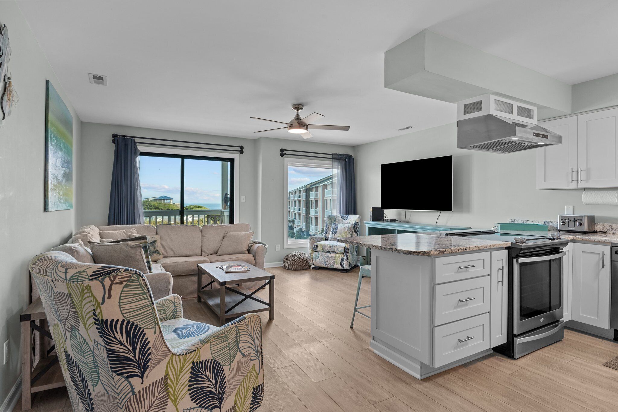Een moderne woonkamer met een bank, een salontafel en een gestreept fauteuil. Er is een keuken met een fornuis, oven en een groot raam.