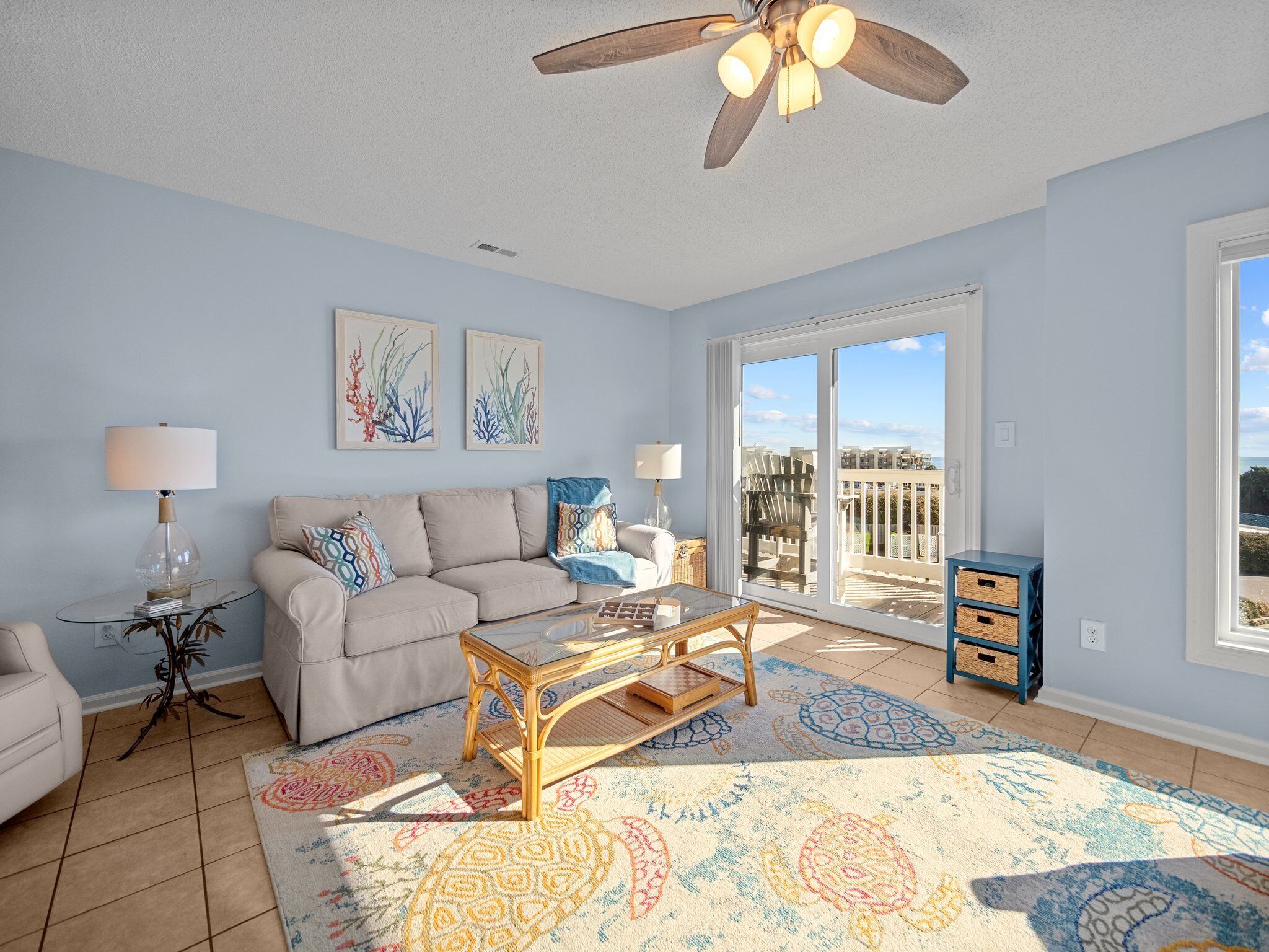 A living room with a sofa, coffee table, and a ceiling fan. There are two windows and a balcony with a view.