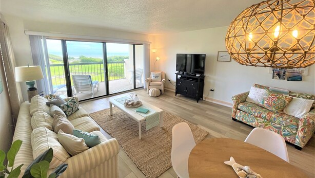 A spacious living room with a large sliding glass door leading to a balcony, a black entertainment unit, a patterned sofa, and a dining area with a table and chairs.
