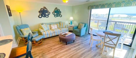 A spacious living room with a wooden floor, a sofa set, a dining table, and a balcony with a view.