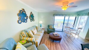 A living room with a sofa, ottoman, and chairs, a ceiling fan, and a view of the ocean through large windows. - Your Slice of Oceanfront Paradise at Queens Court (Emerald Isle)
