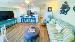 A modern living room with a sofa, ottoman, and a TV on a stand. There is a kitchen area with cabinets and a dining table. - Your Slice of Oceanfront Paradise at Queens Court (Emerald Isle)