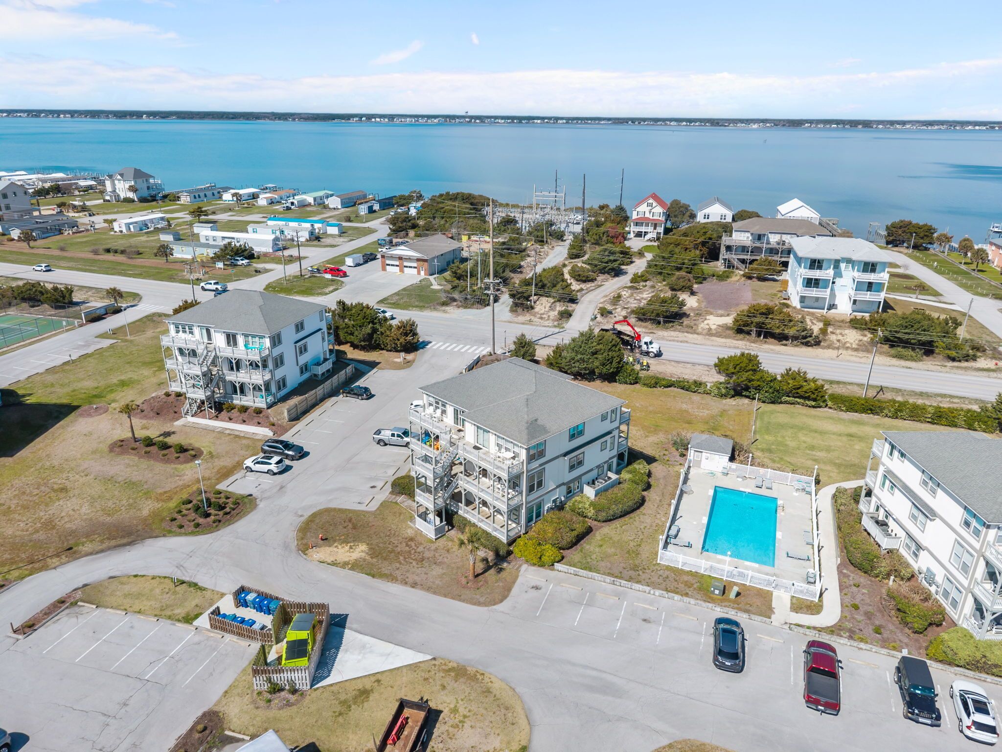 An aerial view of a coastal residential area with multiple buildings, a parking lot, and a large body of water in the background.