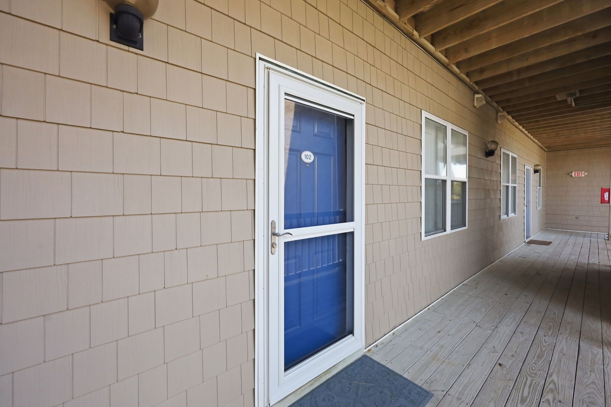 A covered corridor with beige siding, a blue door with the number 102, and a wooden floor.