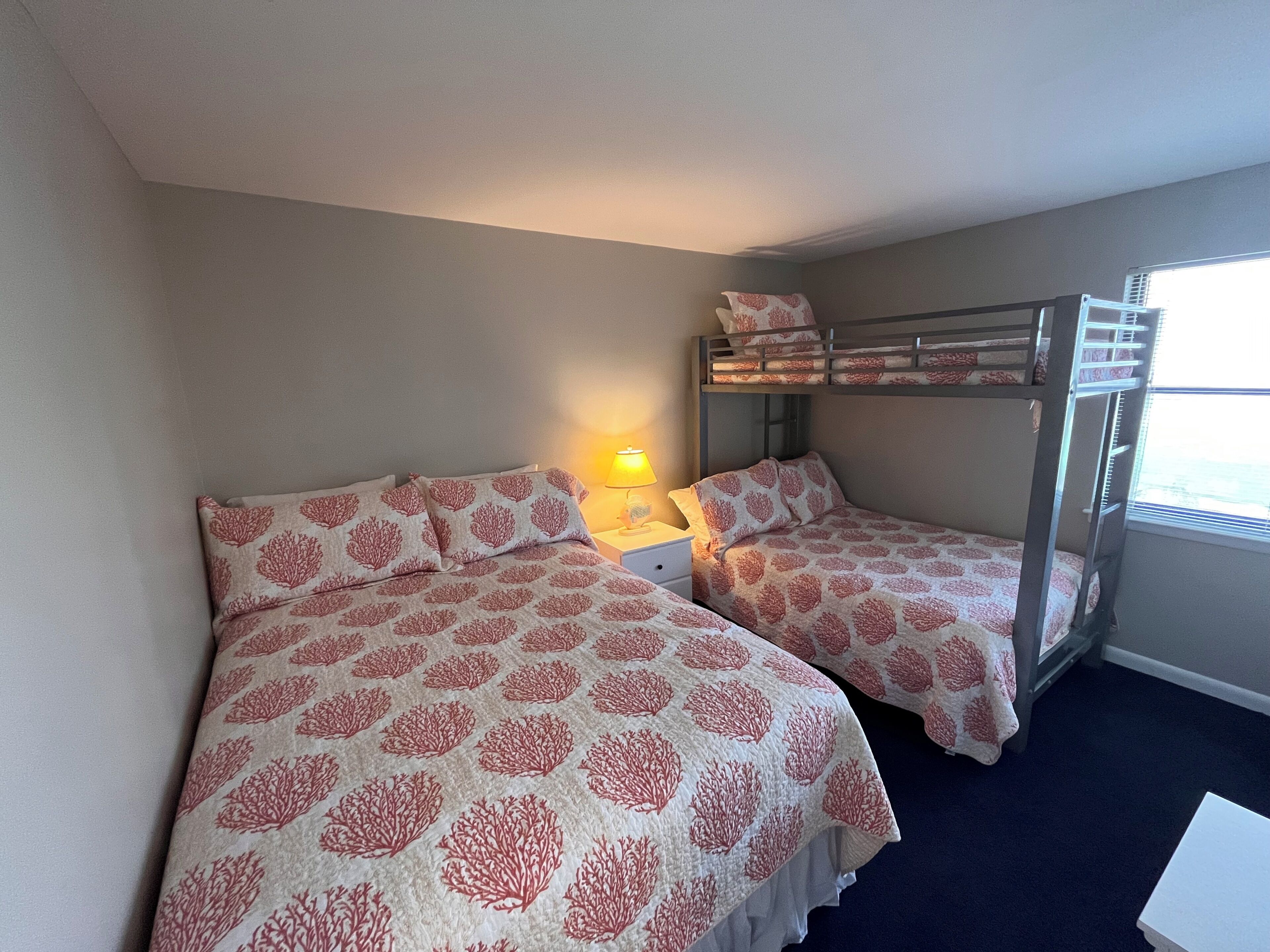A room with two beds, a bunk bed, a bedside lamp, and a window.