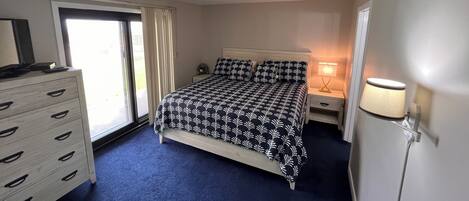 A bedroom with a bed, a dresser, a nightstand, a lamp, and a sliding glass door.