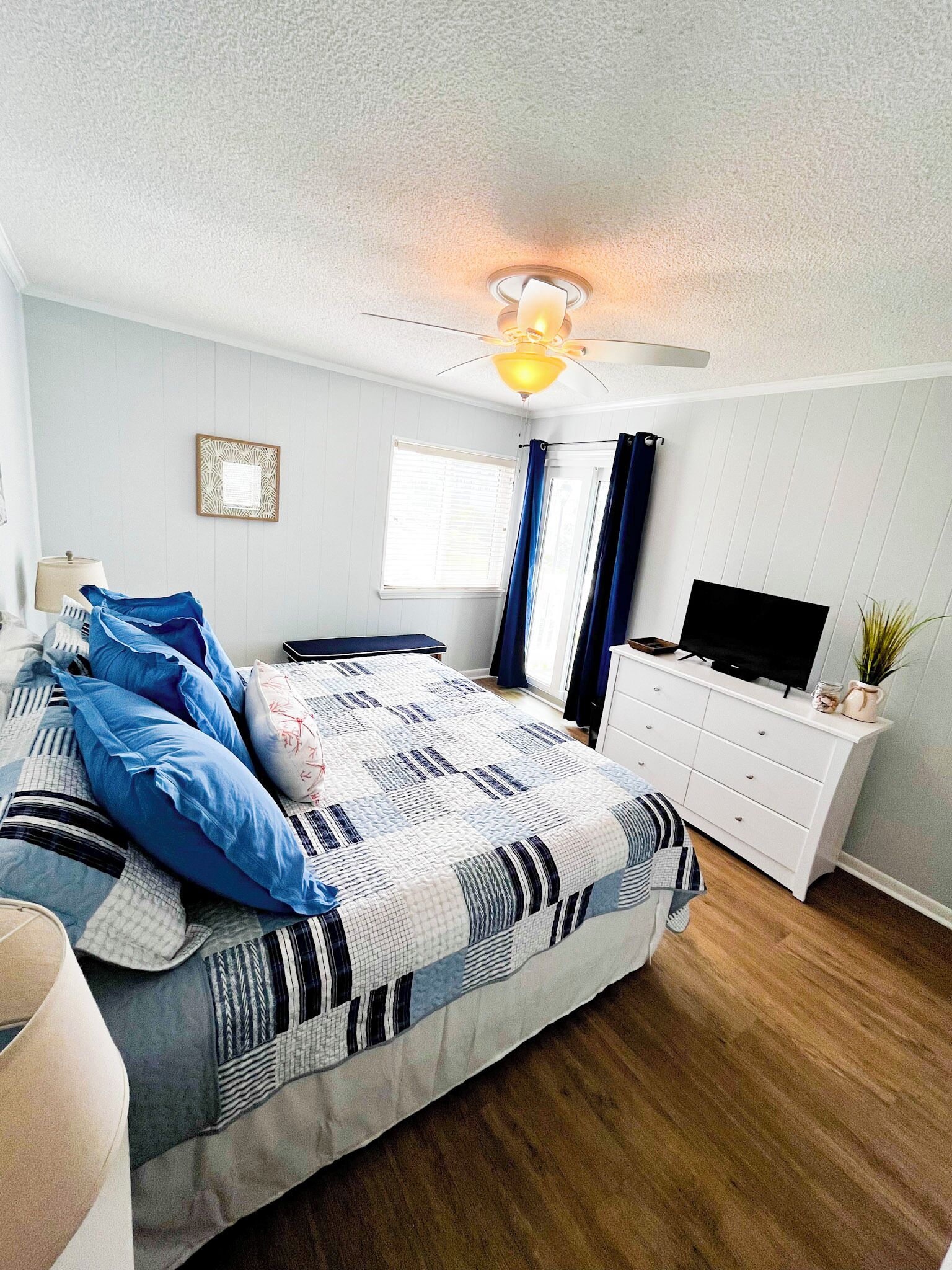 A bedroom with a bed, a TV on a dresser, a ceiling fan, and a window with curtains.