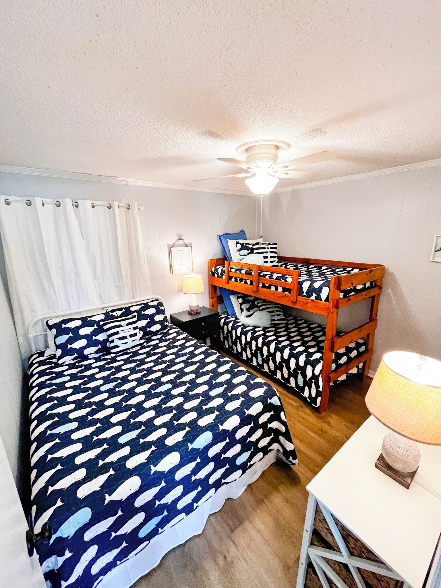 Ein Zimmer mit zwei Betten, eines mit einem gemusterten Bettbezug und das andere mit einem Holzkopfstück. Es gibt einen kleinen Tisch mit einer Lampe, einen Deckenventilator und einen Vorhang am Fenster.