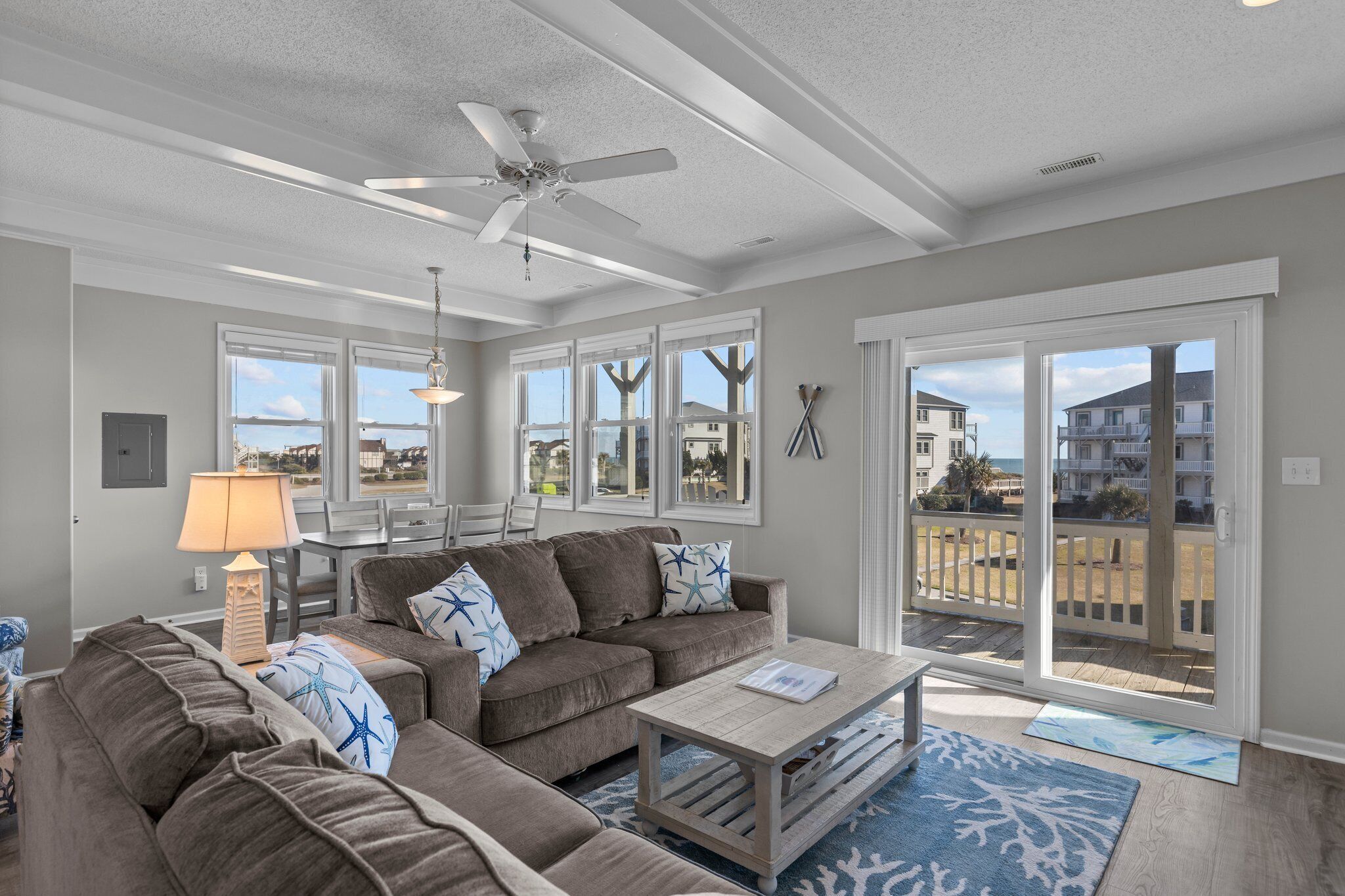 A spacious living room with a ceiling fan, a sofa set, a coffee table, and a view of the beach through large windows.