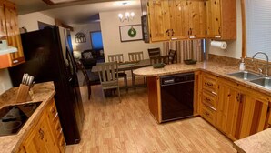 A kitchen with wooden cabinets, a black refrigerator, a dishwasher, and a dining area with chairs and a table. - Cozy Cascade Retreat (Cascade)
