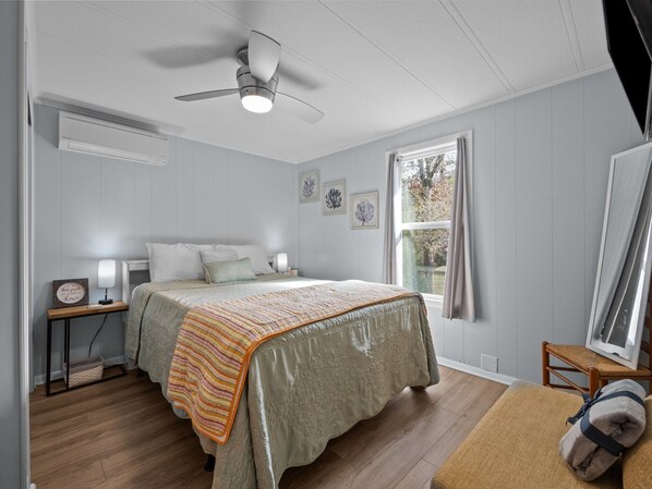 A bedroom with a bed, a ceiling fan, a window, and a nightstand with a lamp. - Duplex with 2 Kitchens | Walk to Beach in Emerald Isle (Emerald isle)