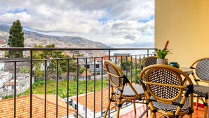 Een balkon met rieten stoelen, een tafel en uitzicht op een stadsgezicht en bergen.