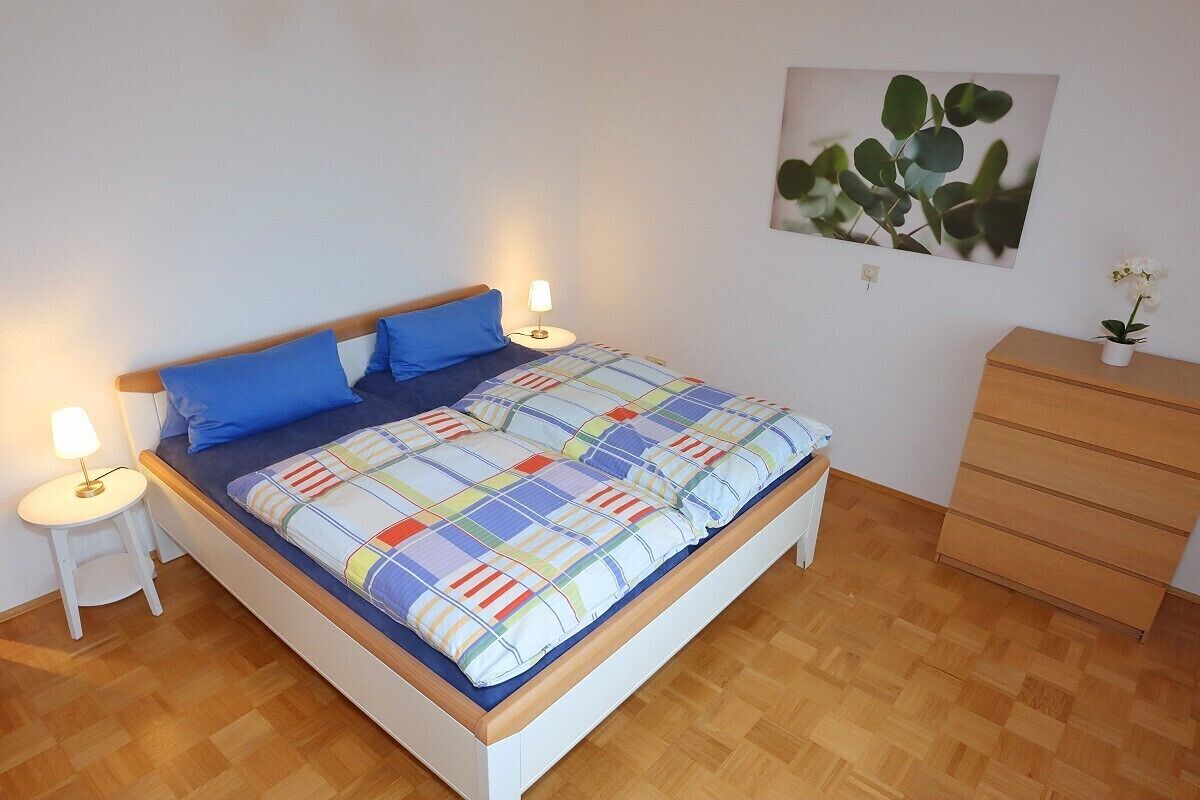 A bed with a colorful quilt, two blue pillows, two bedside lamps, a wooden dresser, and a wall-mounted plant.