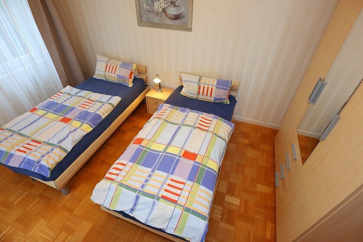 A room with two single beds, each with a colorful quilt and matching pillows. There is a small bedside table with a lamp, and the room has wooden flooring.