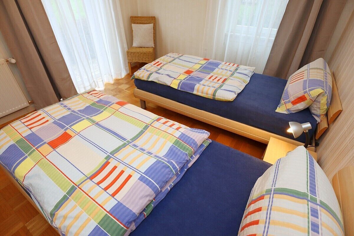A room with two beds, each with a colorful striped bedspread and matching pillows. There is a small table with a lamp, and a chair in the corner.