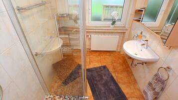 Ein Badezimmer mit Duschkabine aus Glas, einem weißen Waschbecken, einem Handtuchhalter und einem Fenster mit Blick ins Freie.