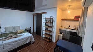 Ein kleines Zimmer mit einem Bett, einem kleinen Schreibtisch, einem Regal mit Pflanzen, einer Tür und einer Küchenzeile.