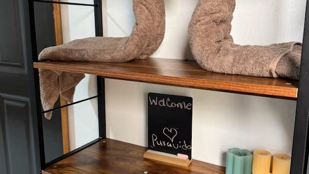 Ein Regal mit zwei schwanenförmigen Handtüchern, einem Kreidetafel-Schild mit 'Welcome Puravida' und Kerzen.