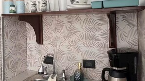Una cocina con un lavamanos de acero inoxidable, una cafetera y una estantería con varios utensilios de cocina.