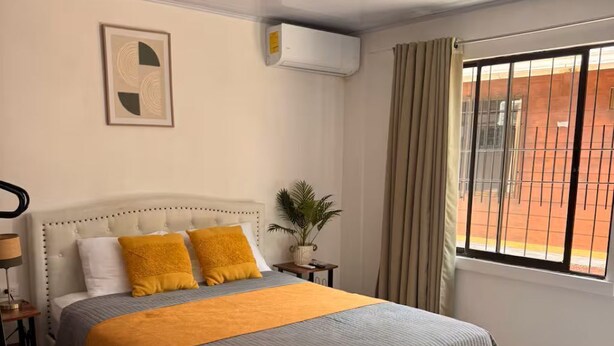 A hotel room with a bed, two orange stools, a potted plant, and a window with curtains. - Cozy Stay in La Fortuna with Parking, WiFi & Smart TV #4 (La Fortuna)