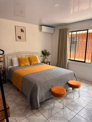 Une chambre d’hôtel avec un lit, deux tabourets orange, une plante en pot et une fenêtre avec des rideaux.