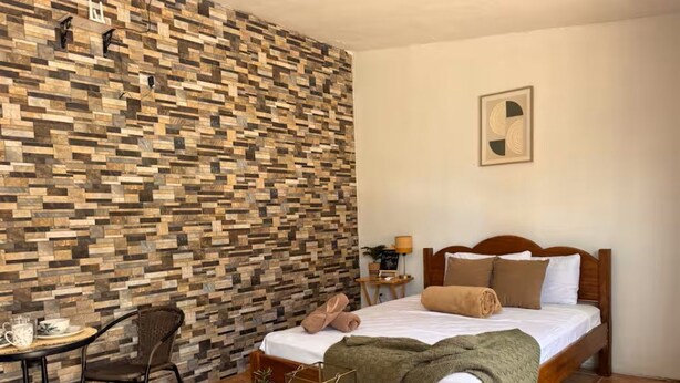 Ett sovrum med en säng i trä, ett litet bord med en stol, en vägg med ett mosaikmönster och en inramad tavla på väggen.