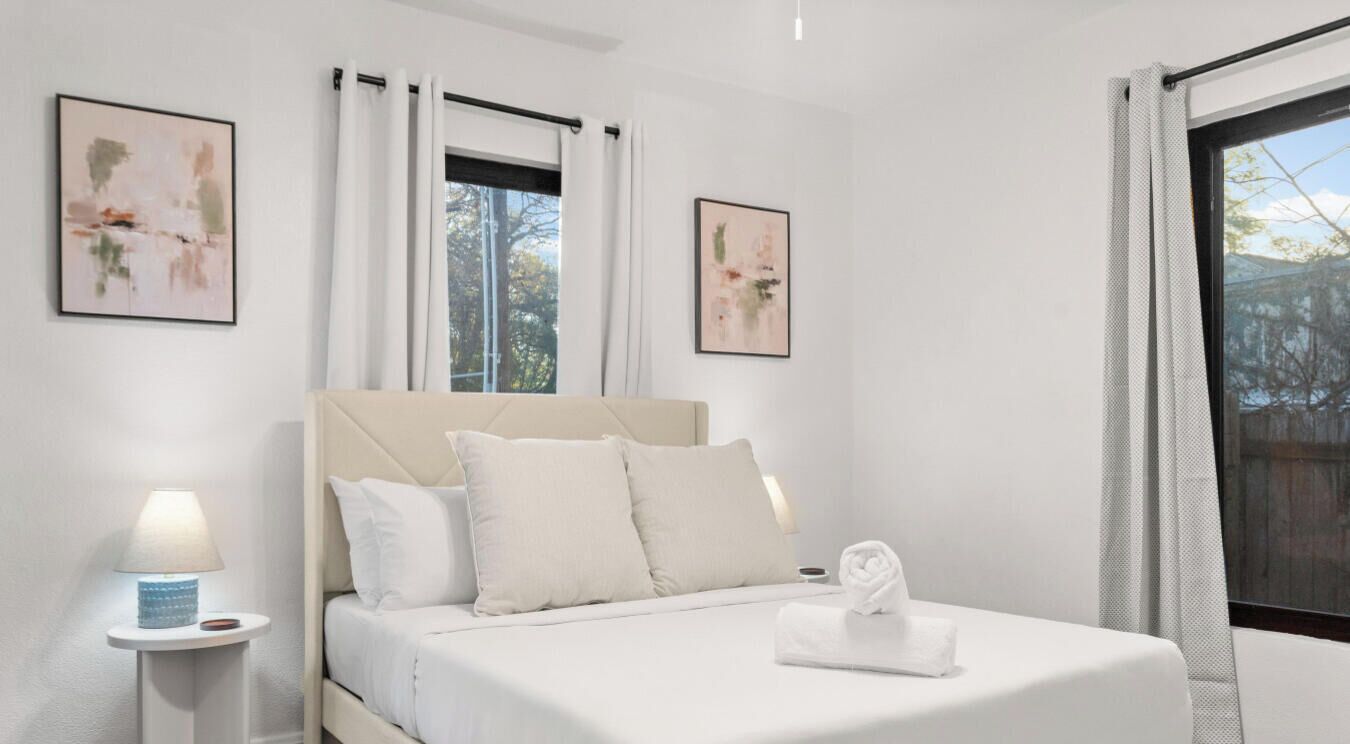 A bedroom with a bed, two windows, a nightstand, and two wall-mounted artworks.
