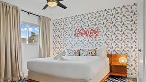 A bedroom with a bed, a ceiling fan, a window with curtains, a wall with a colorful pattern, and a neon sign. - Stylish 1BR/1BA SoCo Condo | Skyline Views, Pool & Parking by Austin Vacay (Austin)
