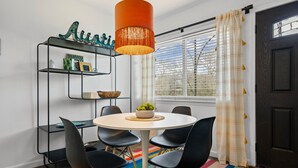 A dining area with a round table, black chairs, a hanging orange pendant light, a metal shelf with decorative items, and a window with blinds. - Stylish 1BR/1BA SoCo Condo | Skyline Views, Pool & Parking by Austin Vacay (Austin)