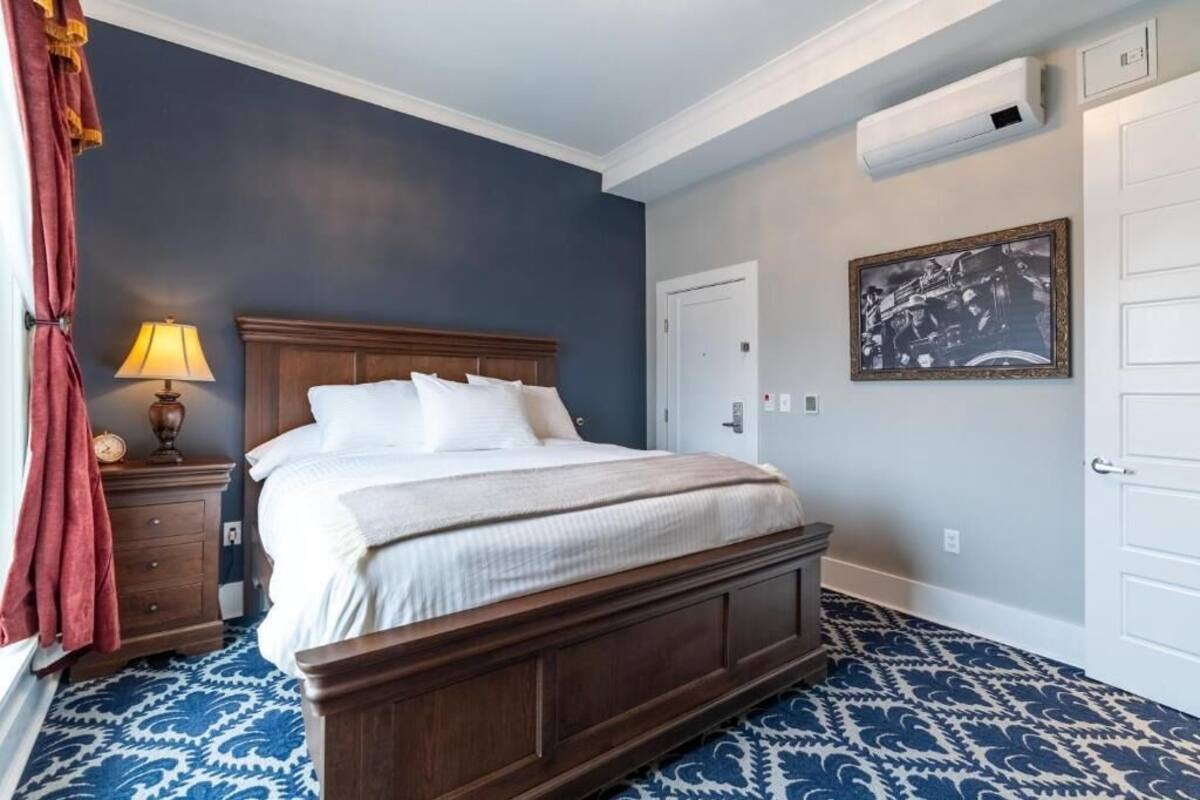 Un dormitorio con una cama de madera, una mesita de noche con una lámpara, una alfombra estampada y un cuadro enmarcado en la pared.