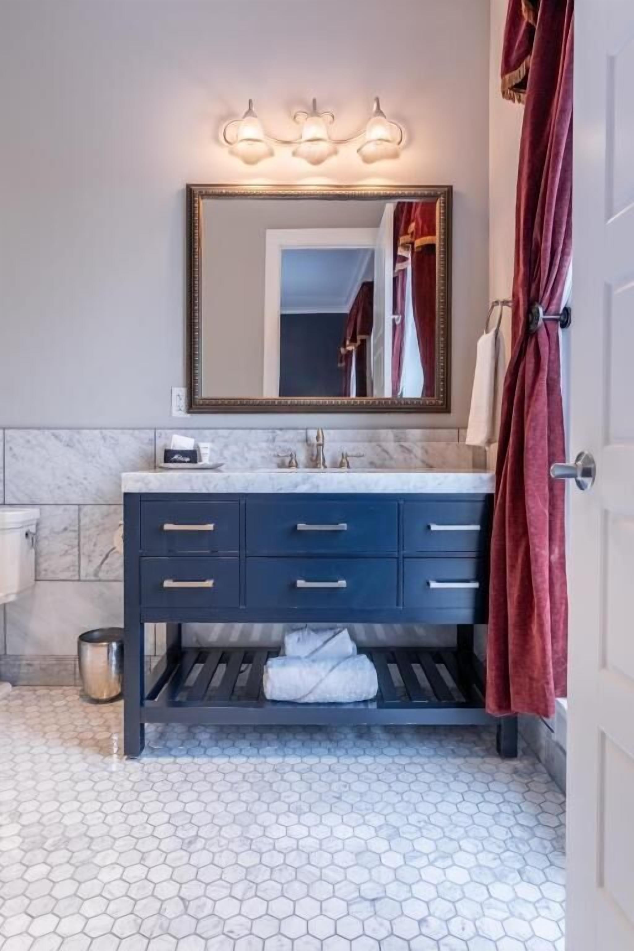 Baño con piso de mármol, tocador oscuro con cajones, espejo con luces y una toalla roja.
