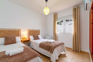A hotel room with two beds, a bedside table with a lamp, a window with curtains, and a hanging light fixture. - Villa Naipa Golf by Villa Plus (Antigua)